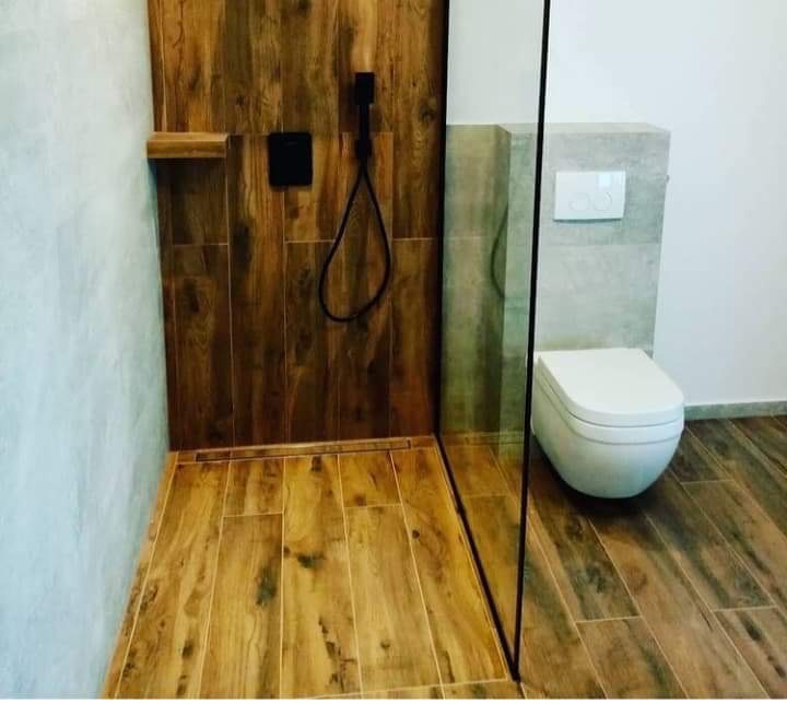 Nowoczesna łazienka z kabiną prysznicową typu walk-in, wykończona płytkami imitującymi drewno, z czarną armaturą i podtynkową spłuczką.