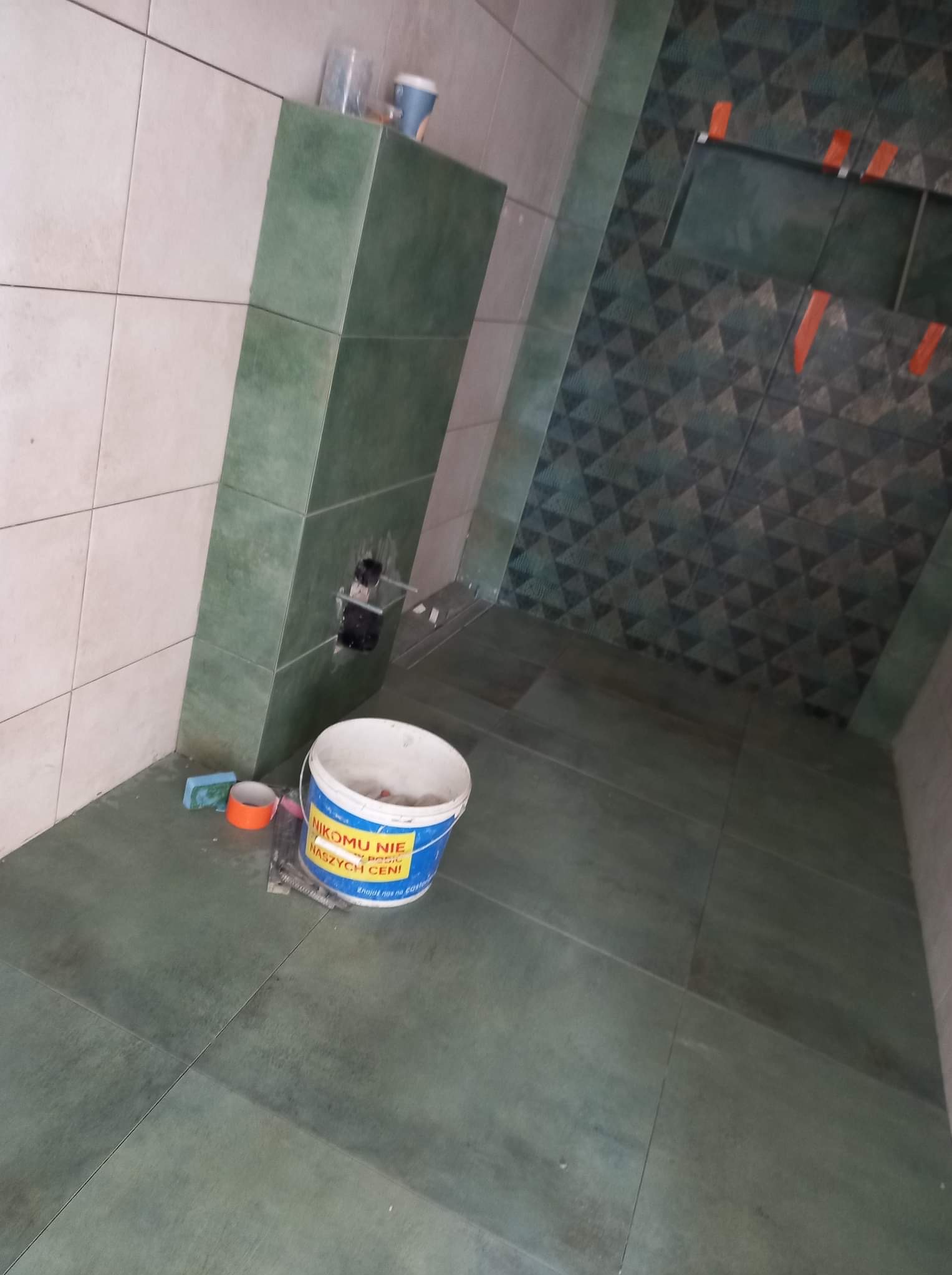 Wnętrze łazienki w trakcie remontu: częściowo wyłożone płytki w odcieniach zieleni i beżu, widoczne narzędzia glazurnicze i wiadro z zaprawą.