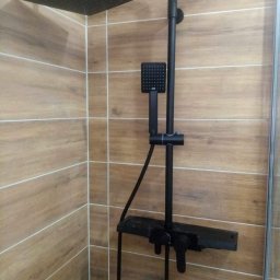 Moric - Czarny zestaw natryskowy z deszczownicą i baterią termostatyczną zamontowany na ścianie wyłożonej płytkami imitującymi drewno.