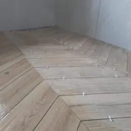 Układanie płytek podłogowych w jodełkę w łazience, widoczne krzyżyki dystansowe, jasne płytki imitujące drewno, szare płytki ścienne w tle.