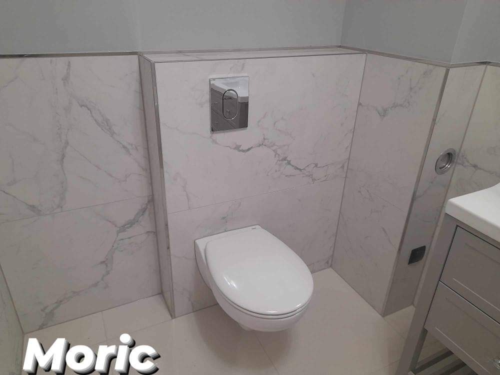 Zawieszona toaleta z płytkami imitującymi marmur i chromowanym przyciskiem spłukującym, wykończona listwami w kolorze aluminium.