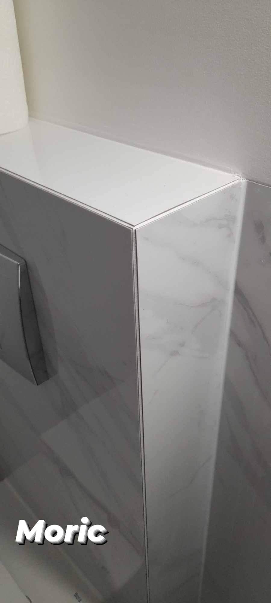 Szczegół narożnika obudowy stelaża WC wykończonego płytkami imitującymi marmur, z widocznym przyciskiem spłukującym i rolką papieru toaletowego.