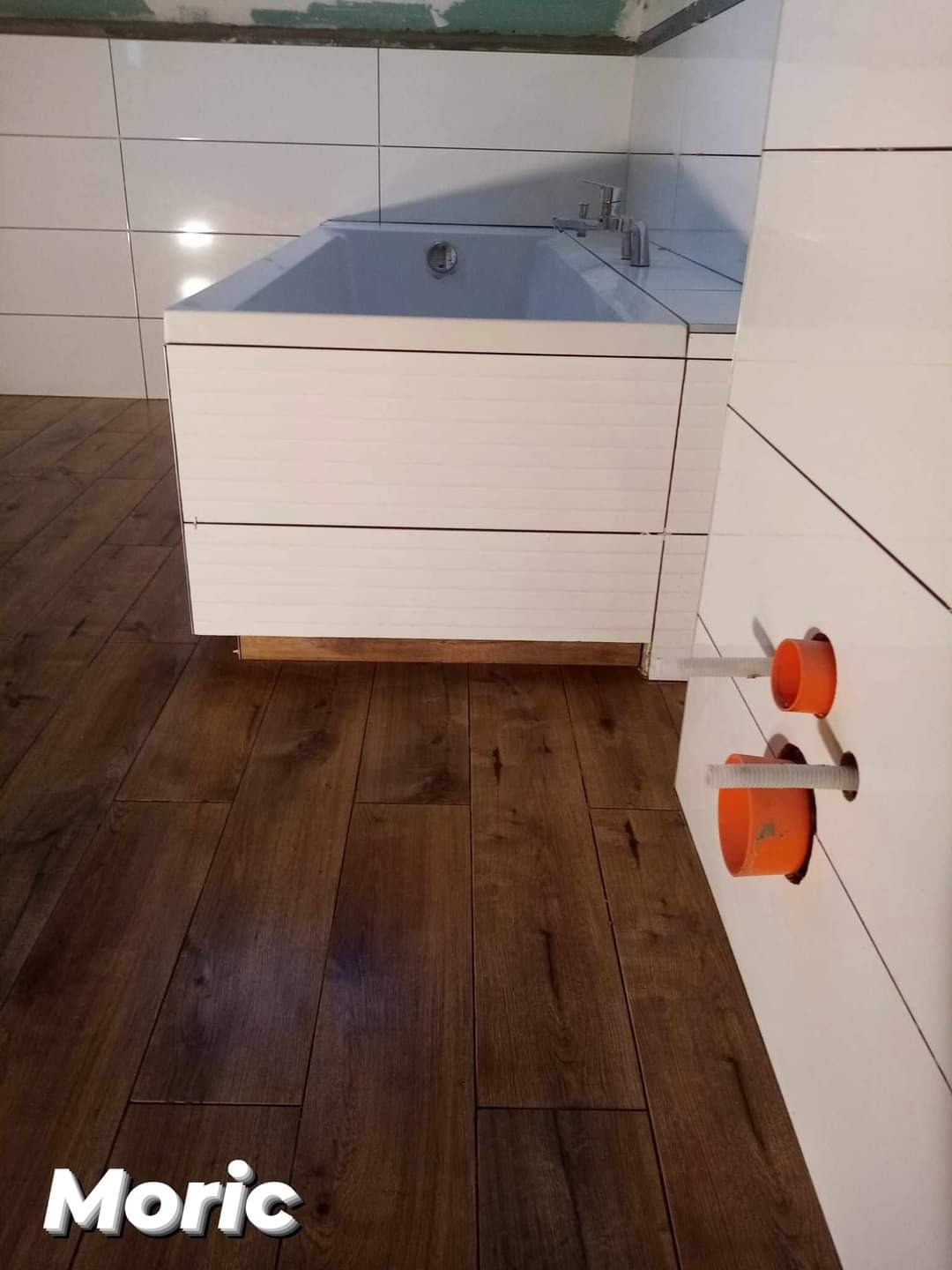 Biała wanna obudowana panelami, jasne płytki na ścianach, ciemna drewnopodobna podłoga, widoczne pomarańczowe elementy instalacji wodnej w ścianie, fragment surowego wykończenia ściany przy suficie.