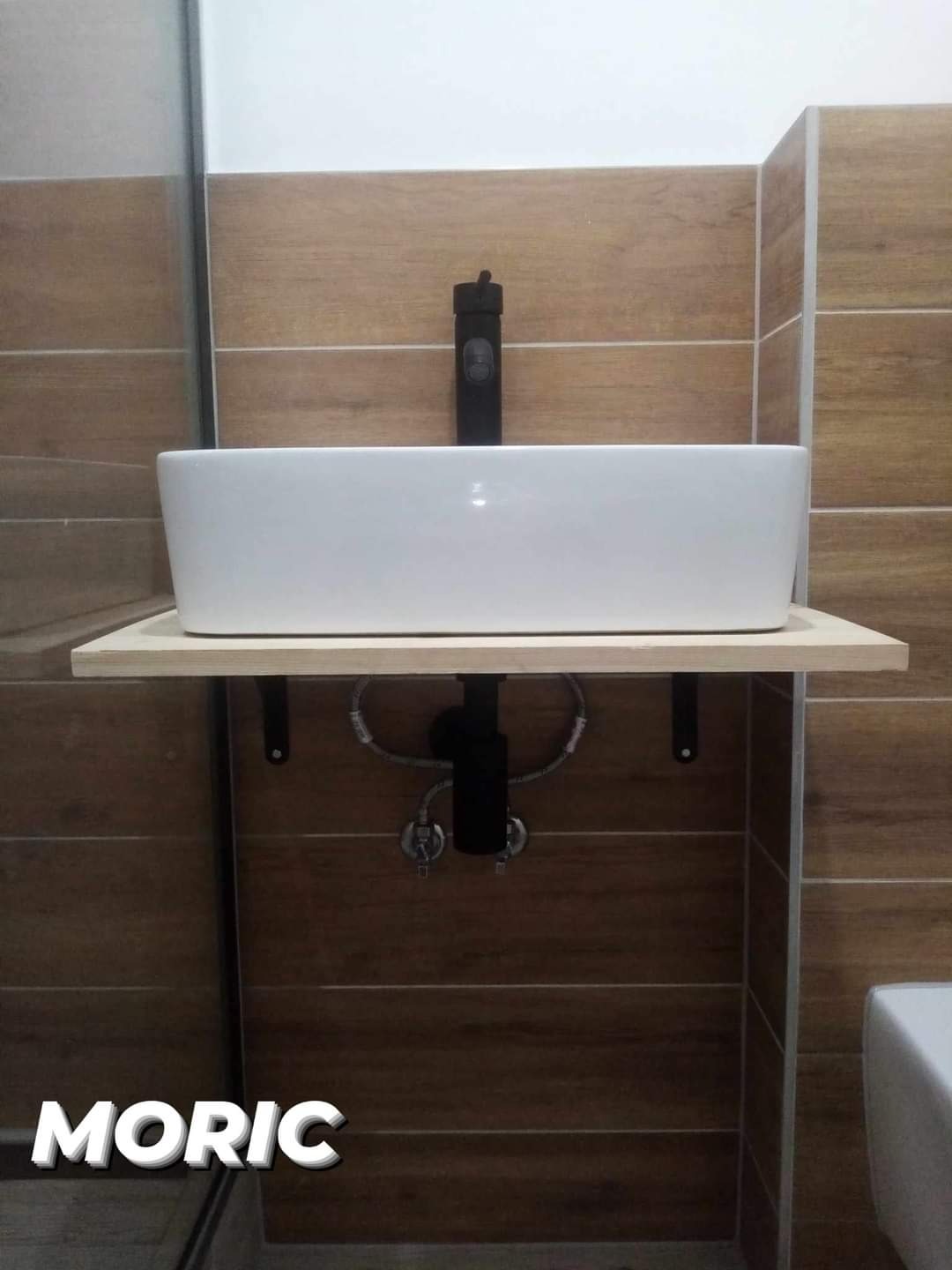 Biała, prostokątna umywalka nablatowa z czarną armaturą na drewnianym blacie, zamontowana na ścianie wyłożonej brązowymi płytkami imitującymi drewno.