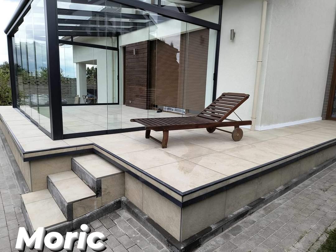 Zadaszony taras z przeszkloną konstrukcją, wyłożony jasnymi płytkami ceramicznymi, z drewnianym leżakiem na kółkach. Schody prowadzą z kostki brukowej na podwyższony poziom tarasu.