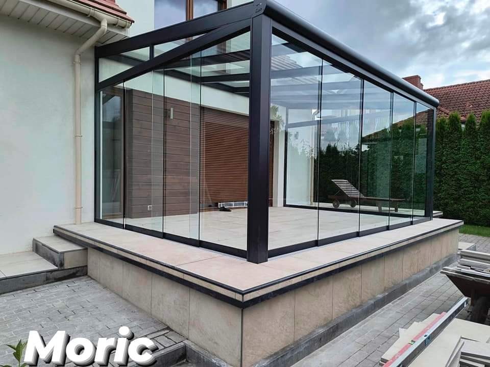 Szklana zabudowa tarasu z czarną ramą, widok z zewnątrz na jasną podłogę i leżak w środku, z boku widoczne schody i fragment ogrodu.