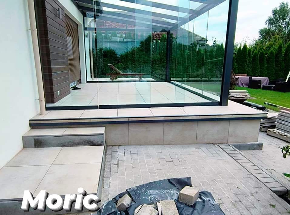 Zadaszony taras z przeszklonymi ścianami i jasnobeżowymi płytkami podłogowymi, widoczne schody wejściowe i przygotowane materiały do dalszych prac wykończeniowych na zewnątrz.