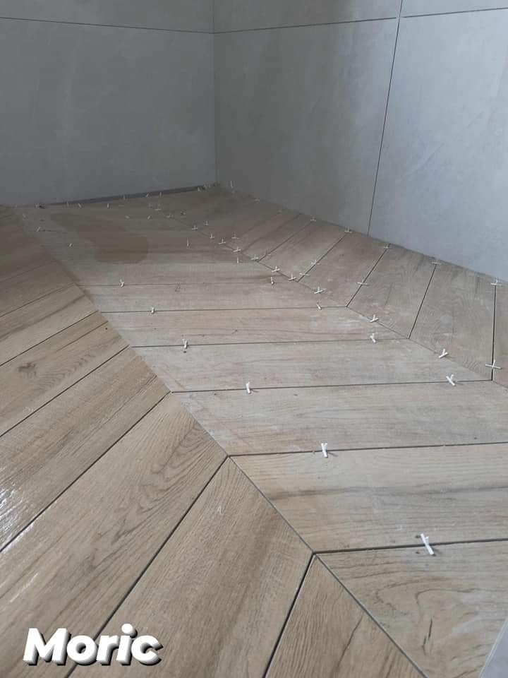 Układanie płytek podłogowych w jodełkę w łazience, widoczne krzyżyki dystansowe, jasne płytki imitujące drewno, szare płytki ścienne w tle.