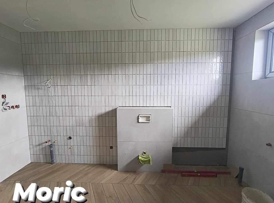 Łazienka w trakcie prac wykończeniowych: ułożone pionowe, białe płytki na ścianach, drewnopodobna podłoga w jodełkę, zabudowa stelaża podtynkowego WC, widoczne instalacje i poziomica.