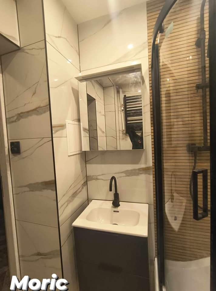 Nowoczesna łazienka z umywalką nablatową na szarej szafce, czarną armaturą i kabiną prysznicową z drewnopodobnymi panelami, wykończona płytkami imitującymi marmur z czarnymi listwami.