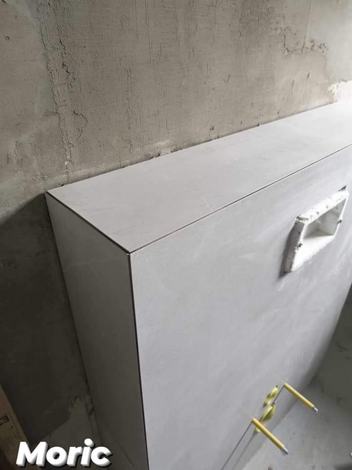 Szara obudowa z płyt gipsowo-kartonowych pokryta białymi płytkami, widoczne żółte krzyżyki dystansowe i otwór na rewizję na tle surowej ściany.