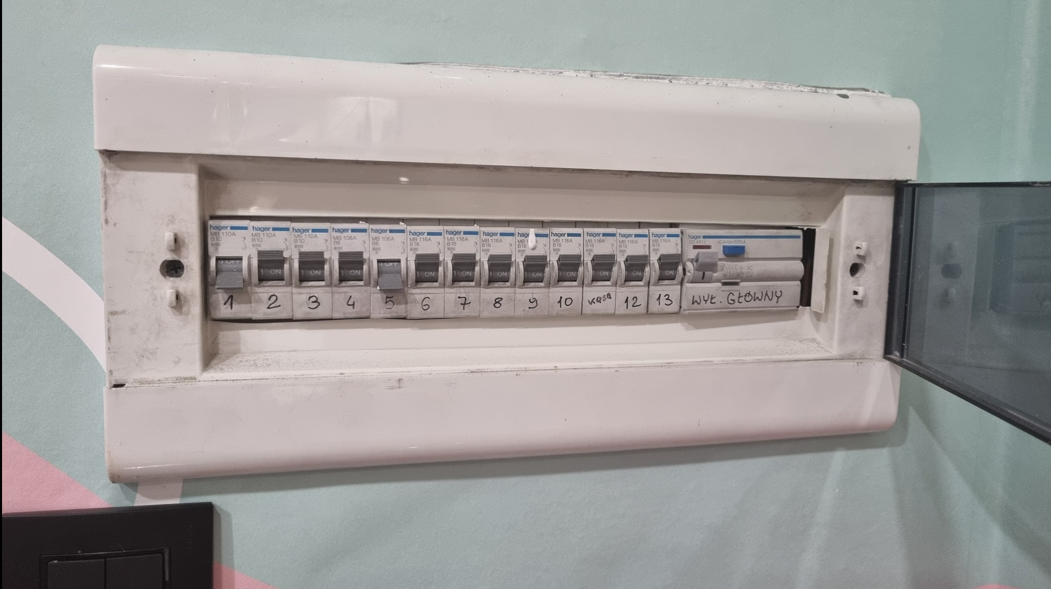 Tablica rozdzielcza z bezpiecznikami Hager, widoczne oznaczenia numerów obwodów i napisy odręczne, częściowo otwarta obudowa, montaż na ścianie z wzorzystą tapetą.