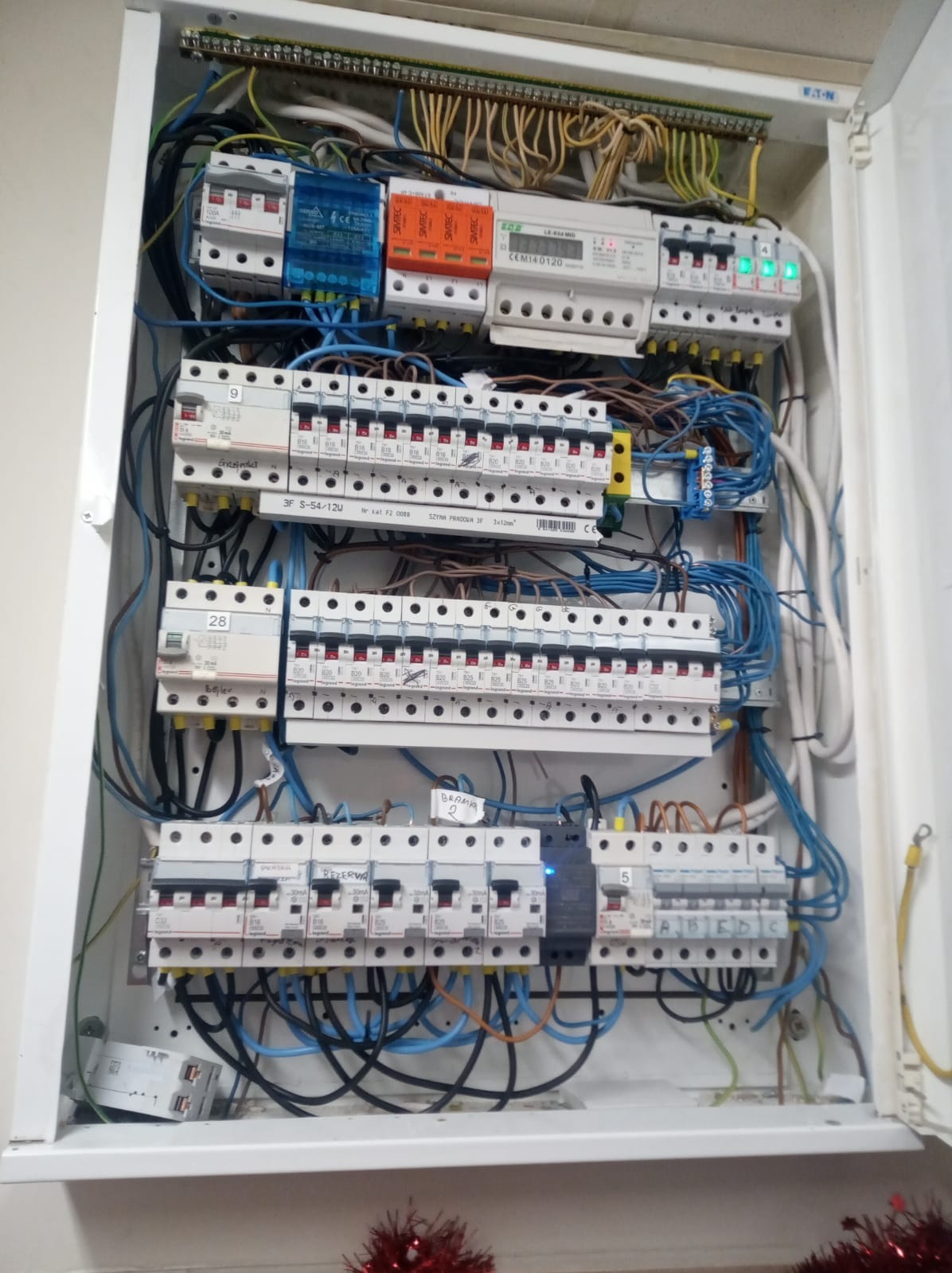 Otwarta skrzynka rozdzielcza z licznymi bezpiecznikami, przewodami w kolorach niebieskim, czarnym, żółtym i brązowym, oraz modułami zabezpieczeń przepięciowych w kolorach niebieskim i pomarańczowym...