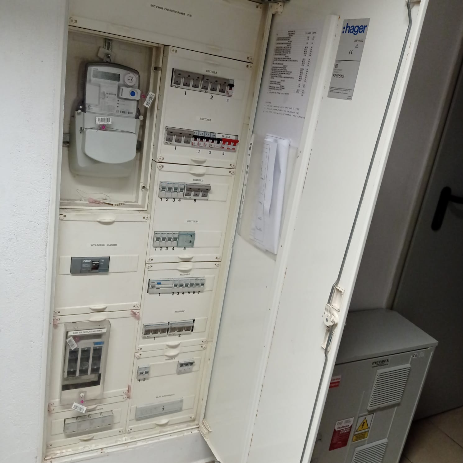Otwarta skrzynka elektryczna z licznikiem i bezpiecznikami, widoczne naklejki informacyjne i oznaczenia na elementach, obok szafka elektryczna Incebex.