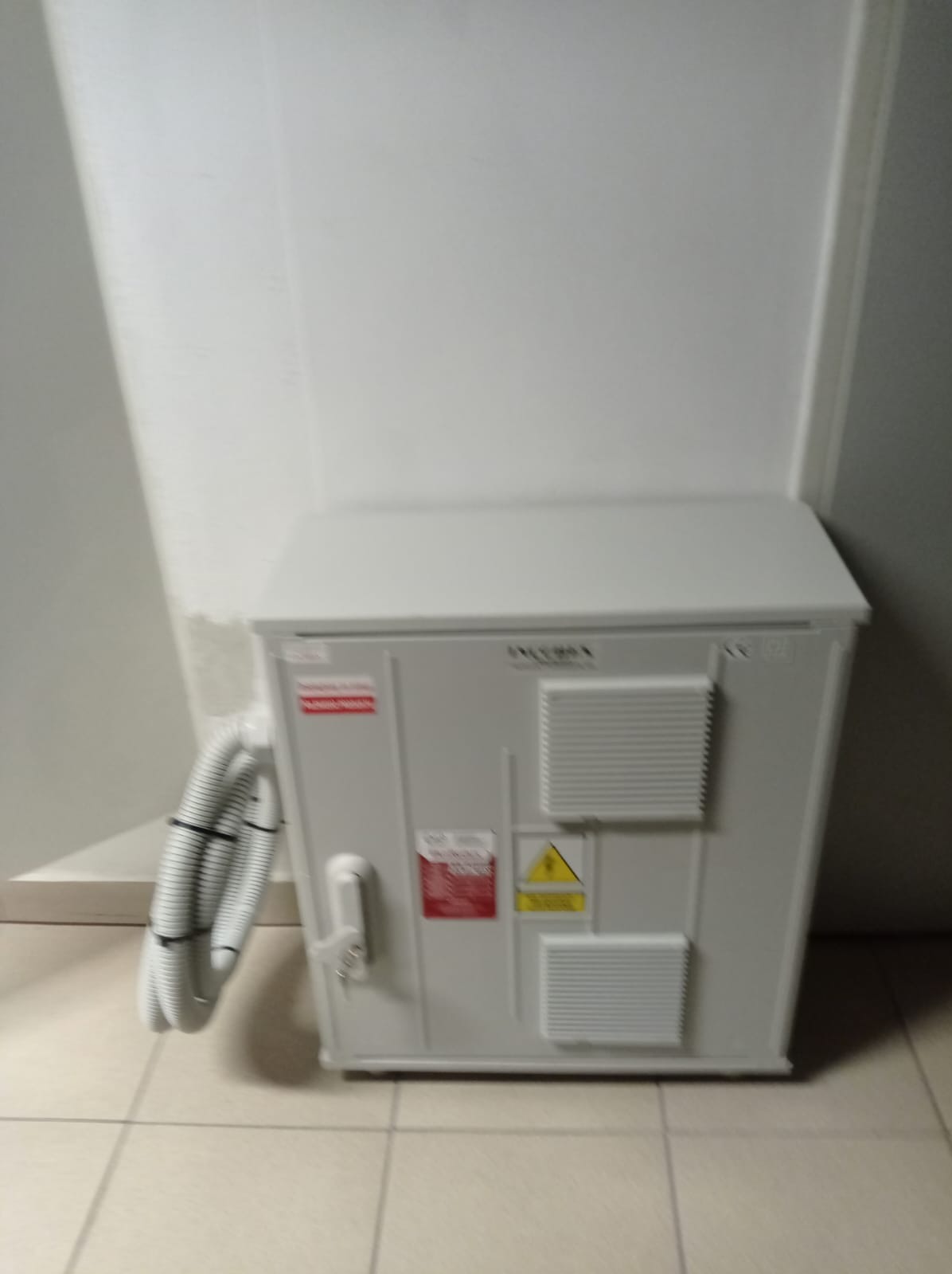 Szara skrzynka elektryczna z widocznymi otworami wentylacyjnymi, naklejkami ostrzegawczymi i przyłączonymi przewodami w białej karbowanej osłonie, umieszczona na podłodze z płytek.