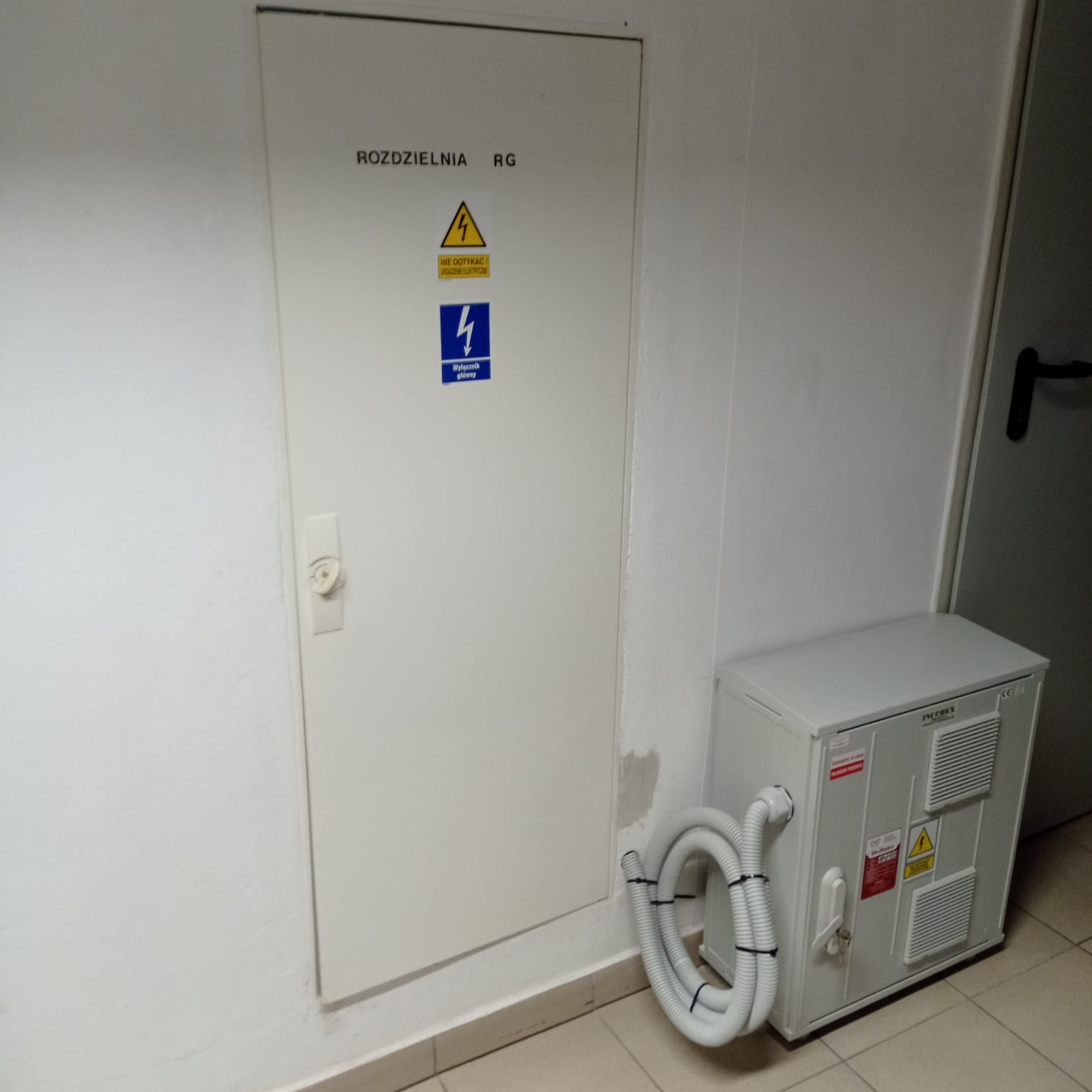 Biała skrzynka rozdzielcza RG z ostrzeżeniami elektrycznymi na ścianie, obok białe urządzenie z podłączonymi rurami wentylacyjnymi.