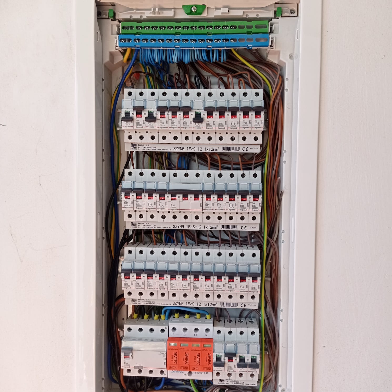Otwarta skrzynka elektryczna z widocznymi bezpiecznikami, przewodami i listwami zaciskowymi, zamontowana na białej ścianie, z etykietami producentów i oznaczeniami technicznymi.