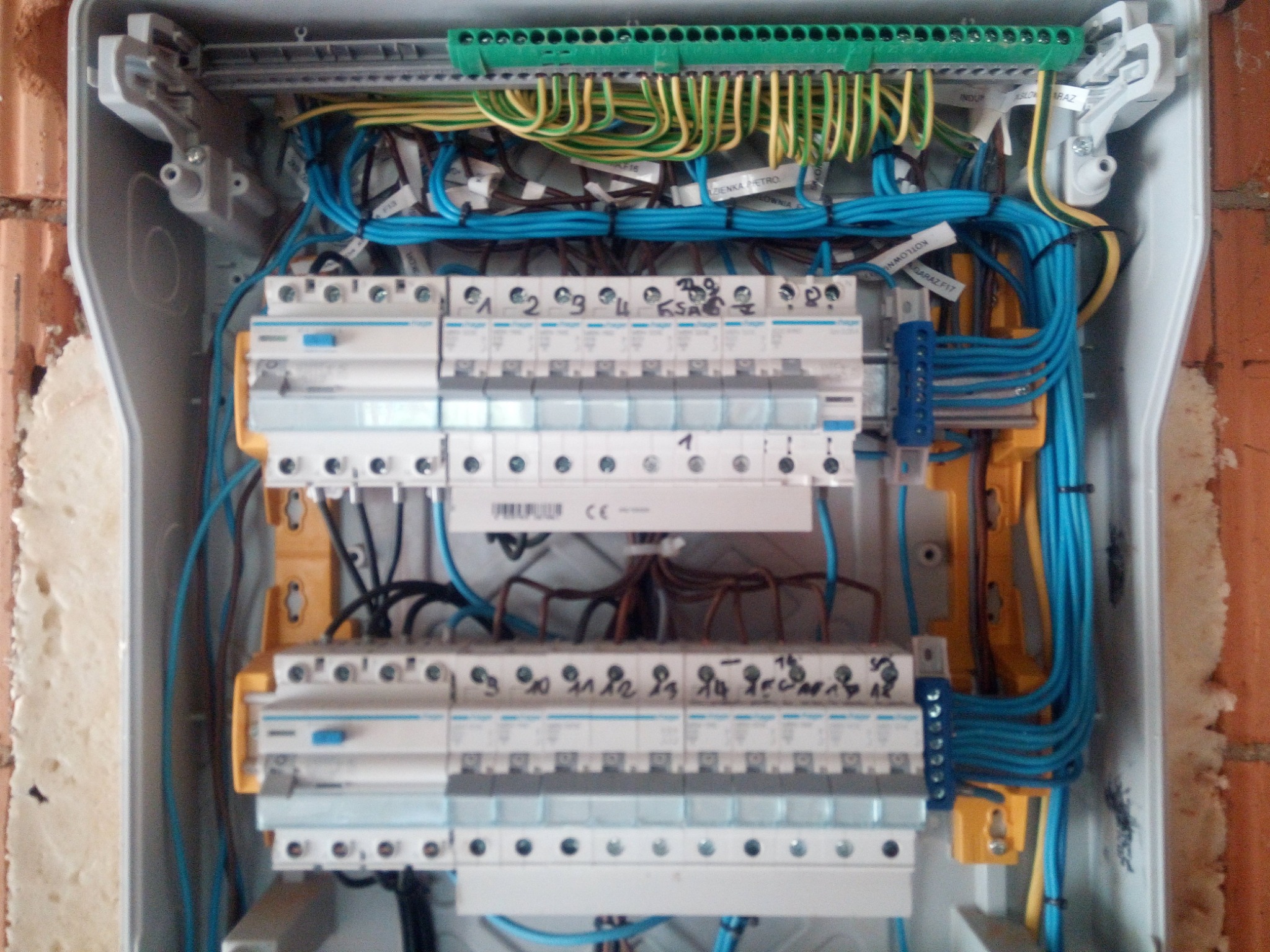 Otwarta skrzynka elektryczna z zamontowanymi bezpiecznikami, okablowaniem w kolorach niebieskim, żółto-zielonym i brązowym, widoczne oznaczenia na przewodach i elementach.
