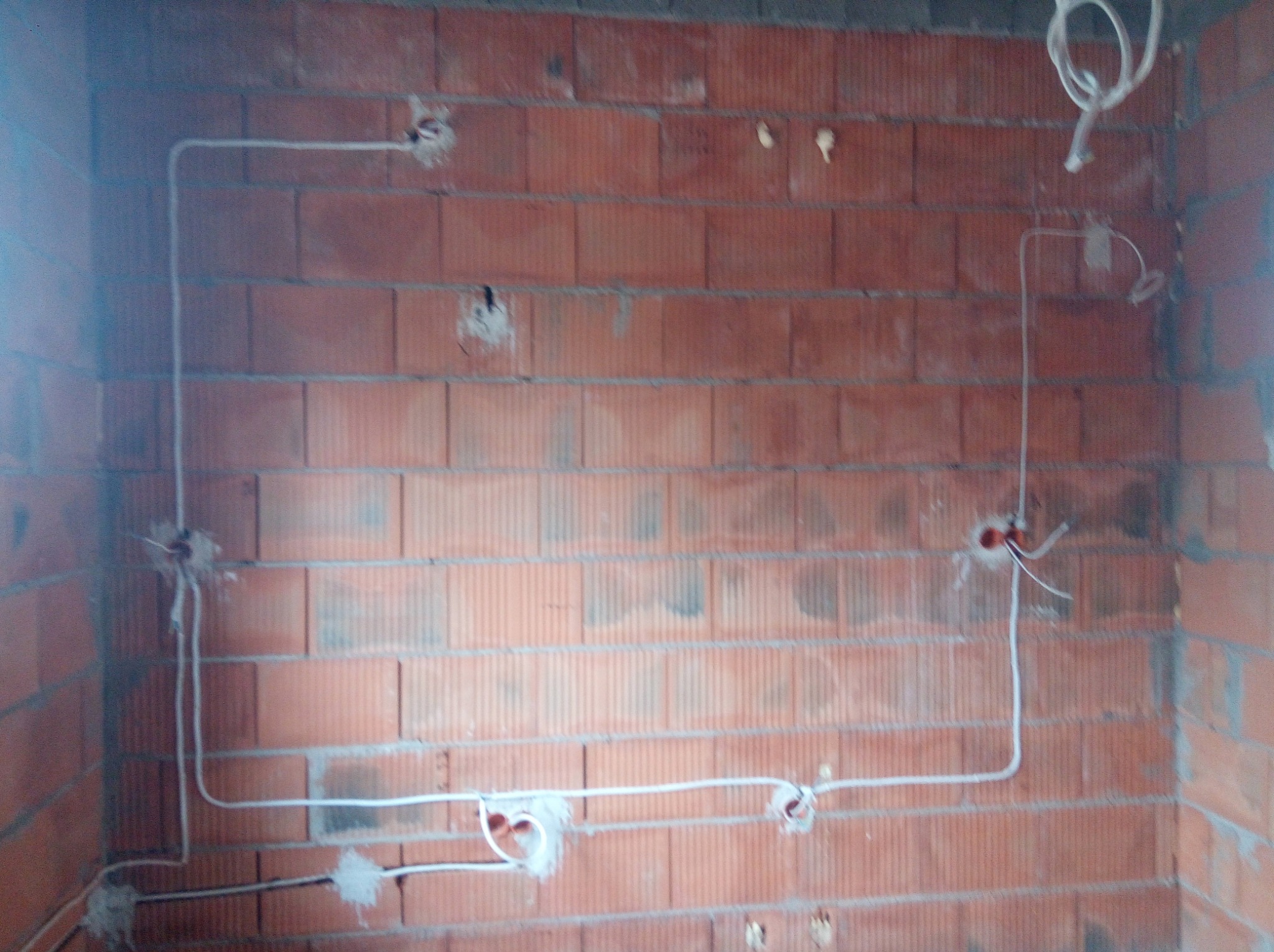 Czerwona ściana z cegły z widocznymi przewodami elektrycznymi poprowadzonymi w bruzdach i gniazdami podtynkowymi, przygotowana do dalszych prac wykończeniowych.