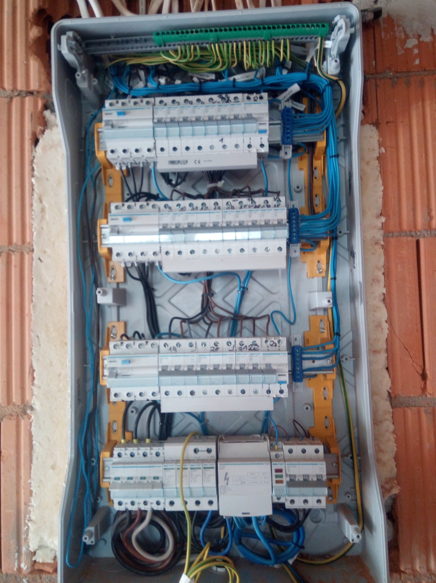 Rozdzielnia elektryczna z bezpiecznikami i okablowaniem, zamontowana na ścianie z cegły w Zgorzelcu, widoczne przewody w kolorach niebieskim, czarnym, żółto-zielonym i brązowym.