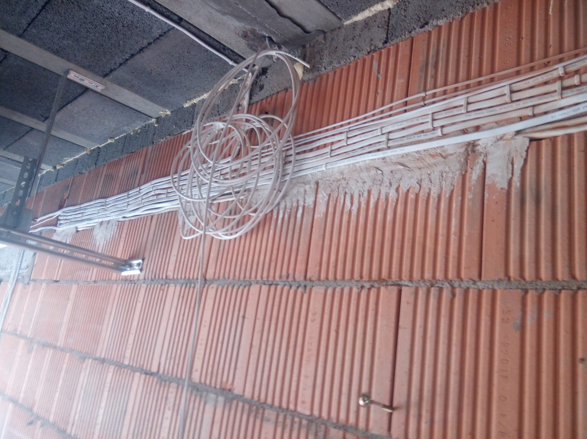 Świeżo ułożone, białe kable elektryczne prowadzone wzdłuż ściany z czerwonej cegły dziurawki, z widocznym mocowaniem do sufitu i zapasem kabla zwisającym w formie pętli.