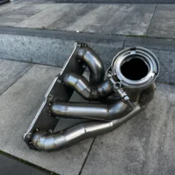 Kolektor wydechowy ze stali nierdzewnej, spawany punktowo, leżący na betonowych schodach, widoczne ślady spawania i montażu.