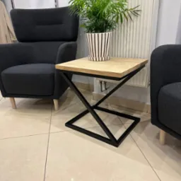 Stolik kawowy z blatem z jasnego drewna i czarną, geometryczną podstawą metalową, obok szarych foteli i grzejnika.