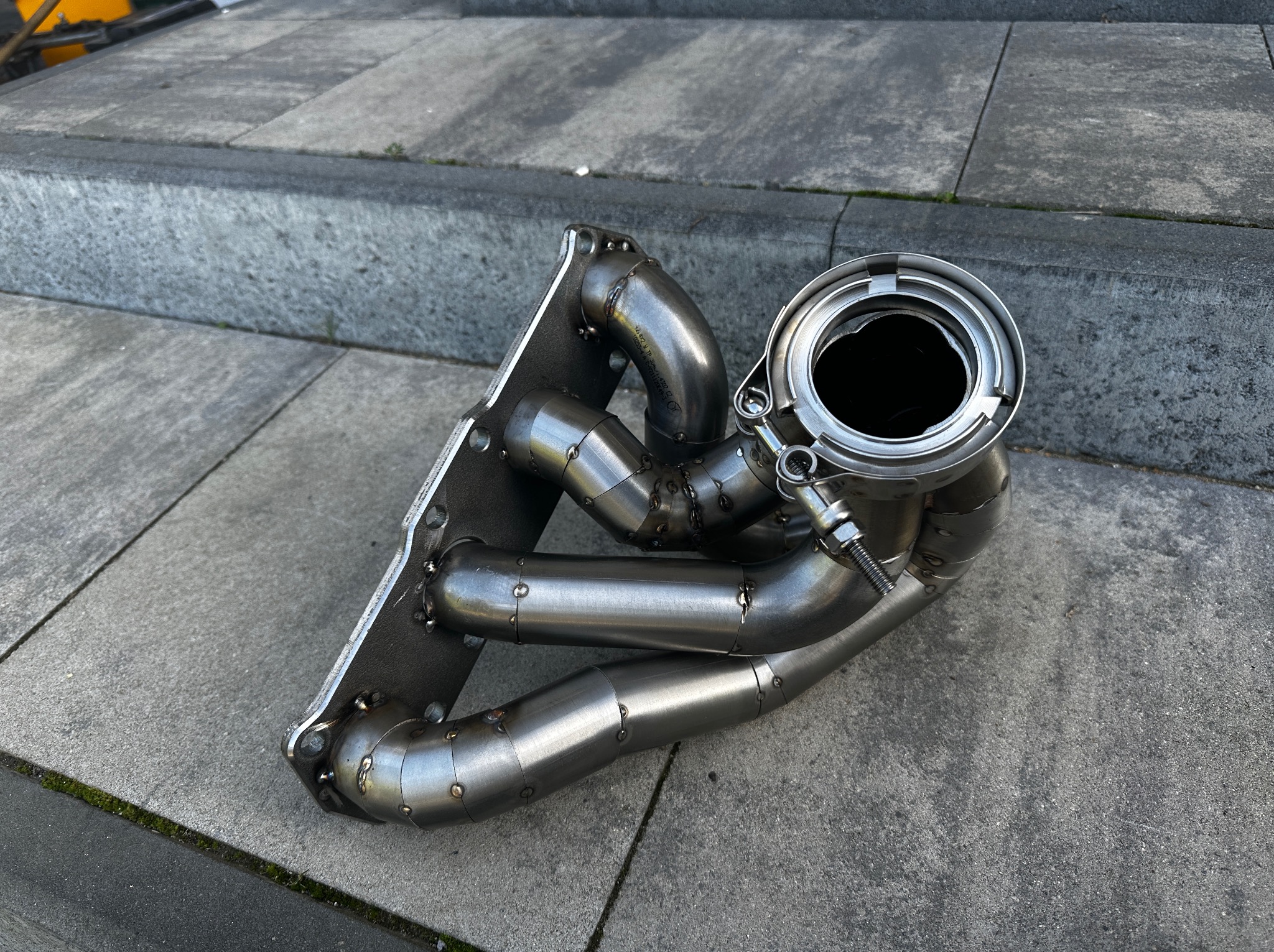 Kolektor wydechowy ze stali nierdzewnej, spawany punktowo, leżący na betonowych schodach, widoczne ślady spawania i montażu.