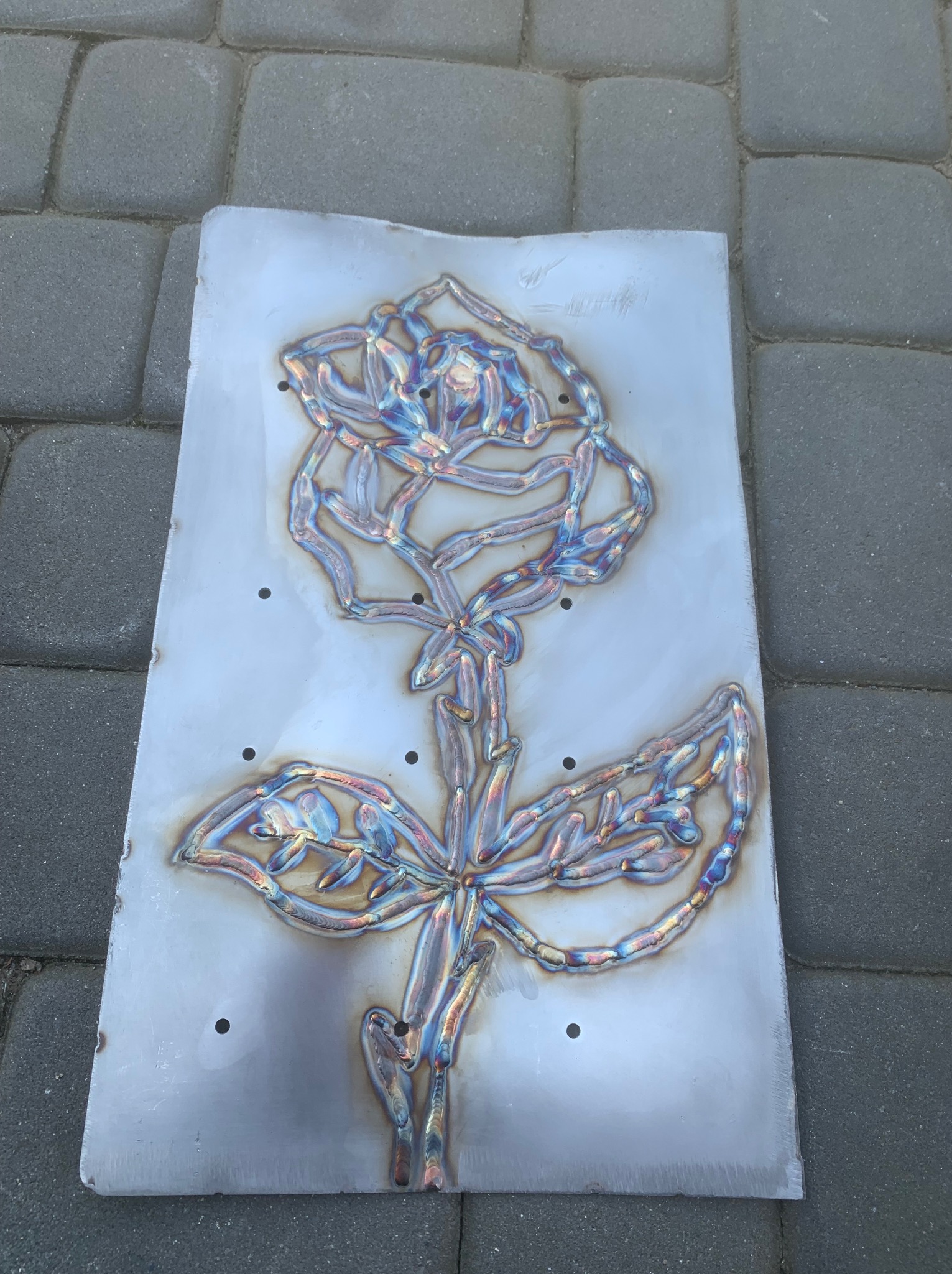 Metalowa płyta z wyciętym wzorem róży za pomocą spawania, widoczne ślady termiczne i otwory montażowe, leżąca na brukowanej powierzchni.