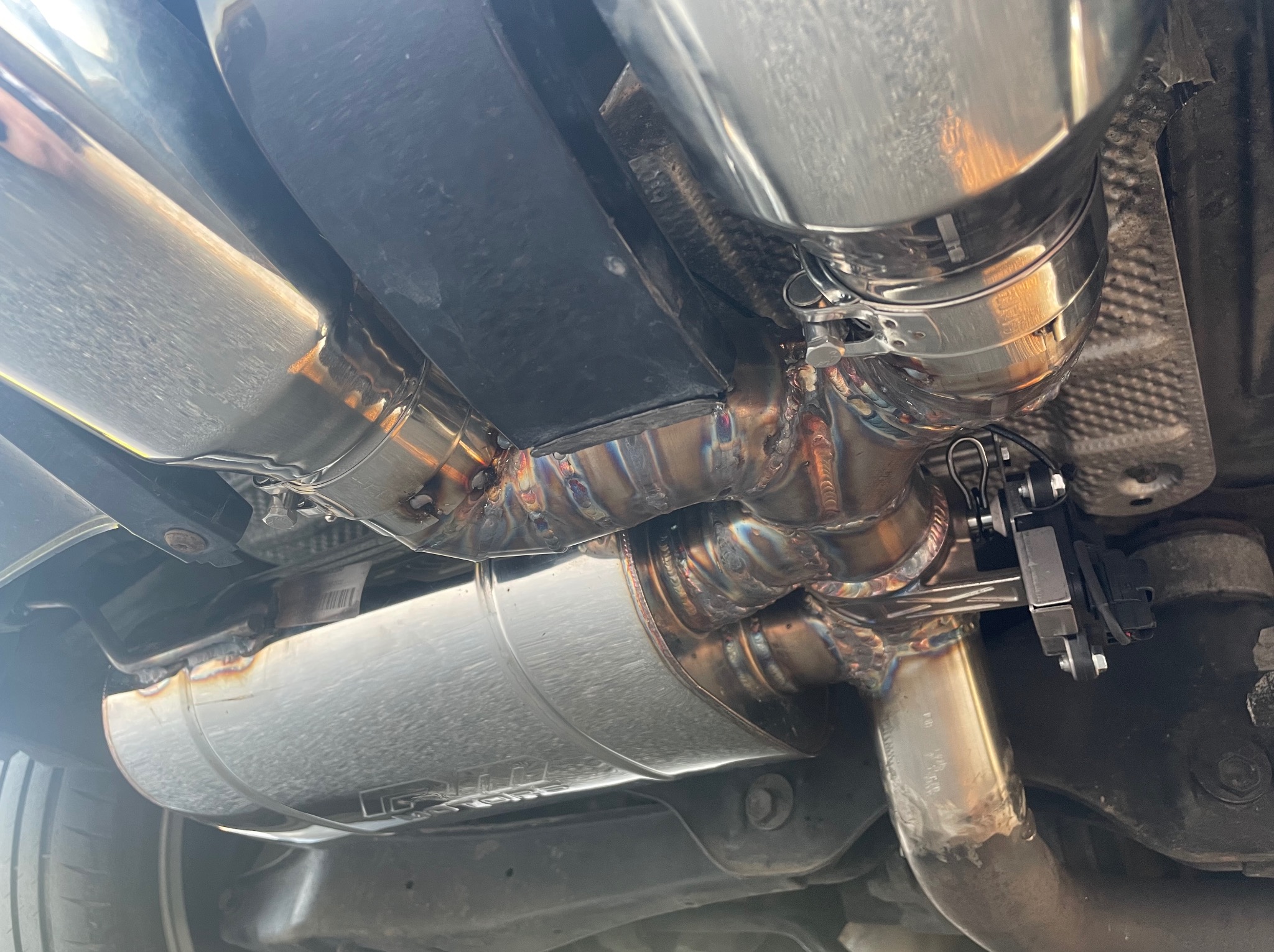 Widok od spodu na zamontowany, spawany, nierdzewny układ wydechowy samochodu z widocznymi spawami i elementami mocującymi.