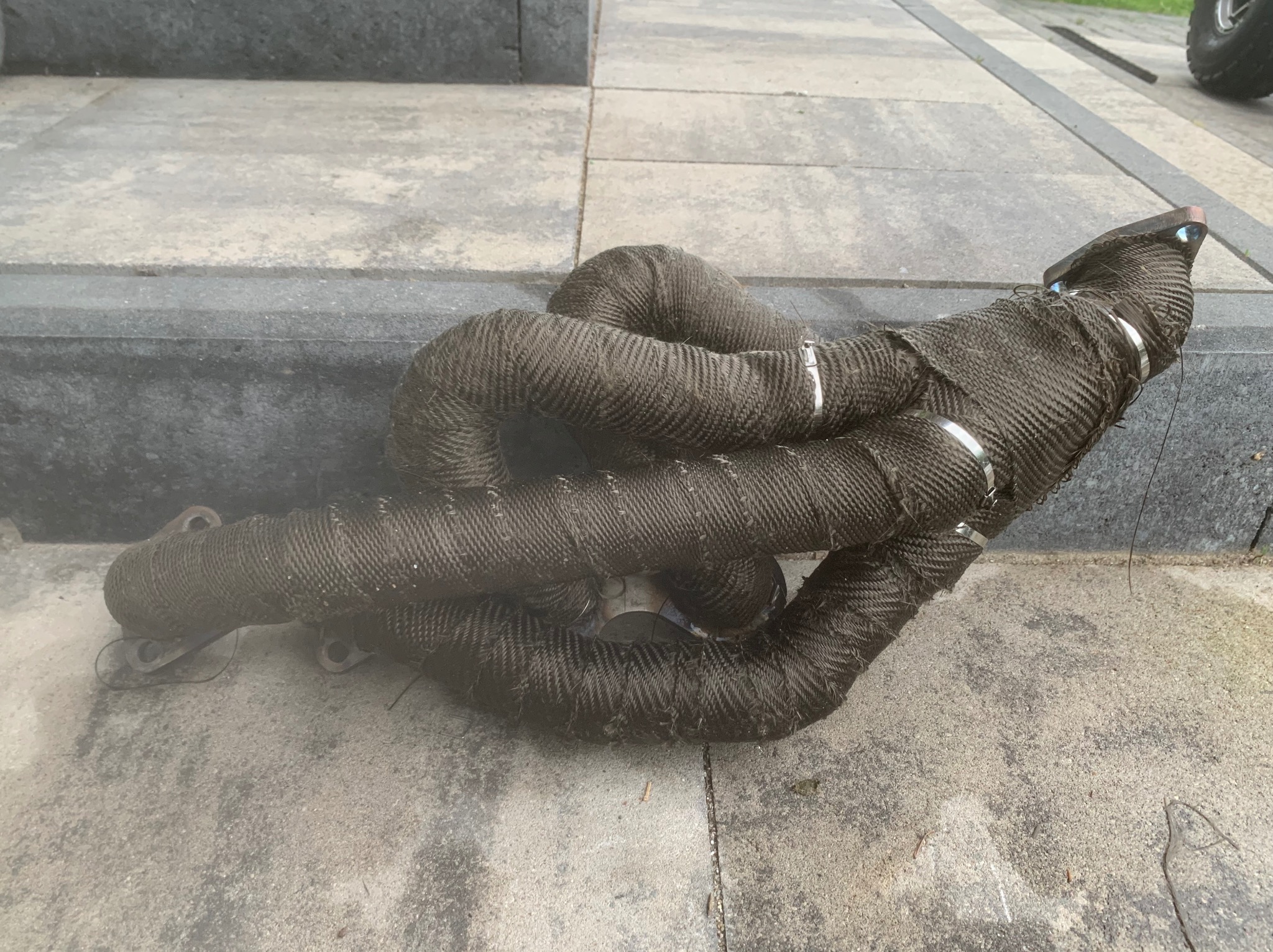 Kolektor wydechowy owinięty termoizolacyjną taśmą na betonowej nawierzchni, zabezpieczony metalowymi opaskami zaciskowymi.