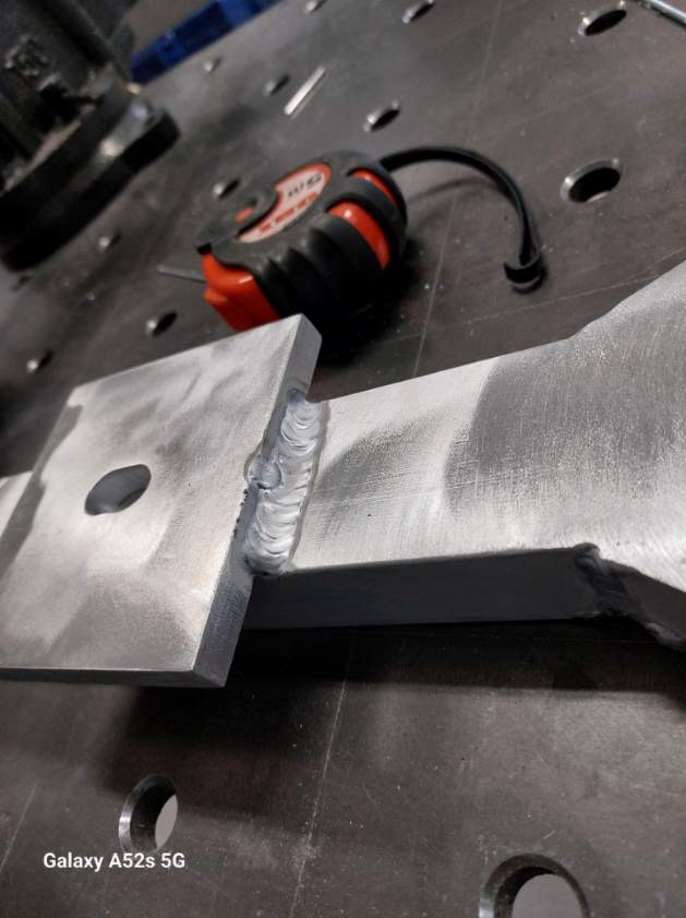 Spoina na metalowej konstrukcji z widocznym szwem spawalniczym, leżąca na metalowym stole spawalniczym obok zwiniętej miarki.