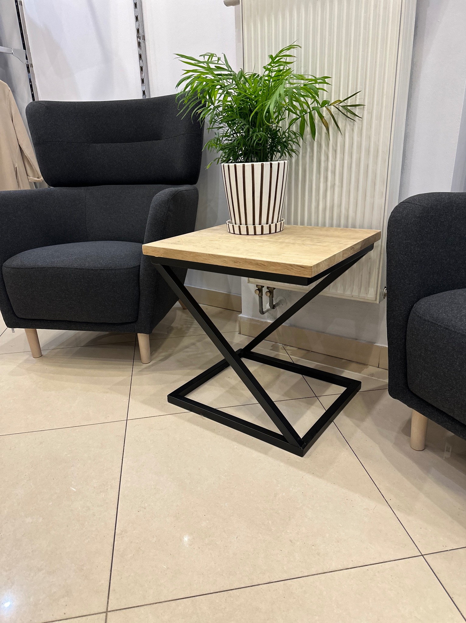 Stolik kawowy z blatem z jasnego drewna i czarną, geometryczną podstawą metalową, obok szarych foteli i grzejnika.