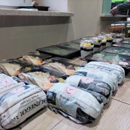 Bobrovsky catering - Zestawy cateringowe: sałatki w czarnych pojemnikach, desery z owocami w przezroczystych kubkach i kanapki zawinięte w papier z nadrukowanym tekstem, ułożone rzędami na drewnianym blacie.