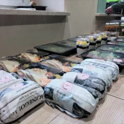 Zestawy cateringowe: sałatki w czarnych pojemnikach, desery z owocami w przezroczystych kubkach i kanapki zawinięte w papier z nadrukowanym tekstem, ułożone rzędami na drewnianym blacie.