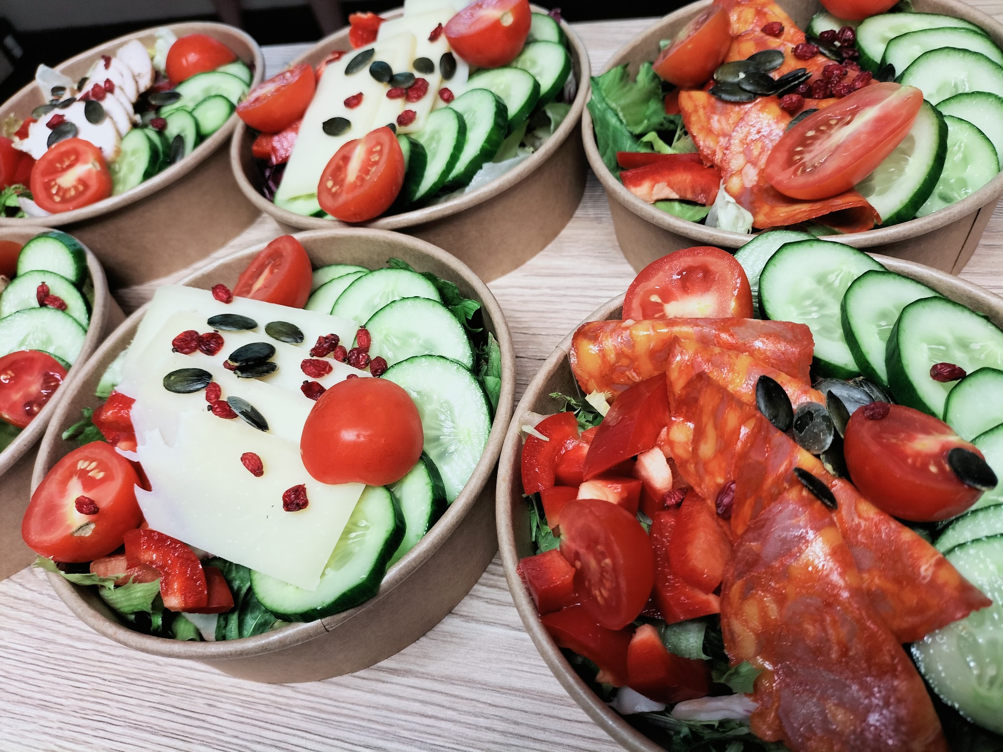 Różnorodne sałatki w papierowych miskach: jedna z plastrami sera i pomidorami, druga z pikantnym salami, ogórkami i pestkami dyni, na drewnianym blacie.