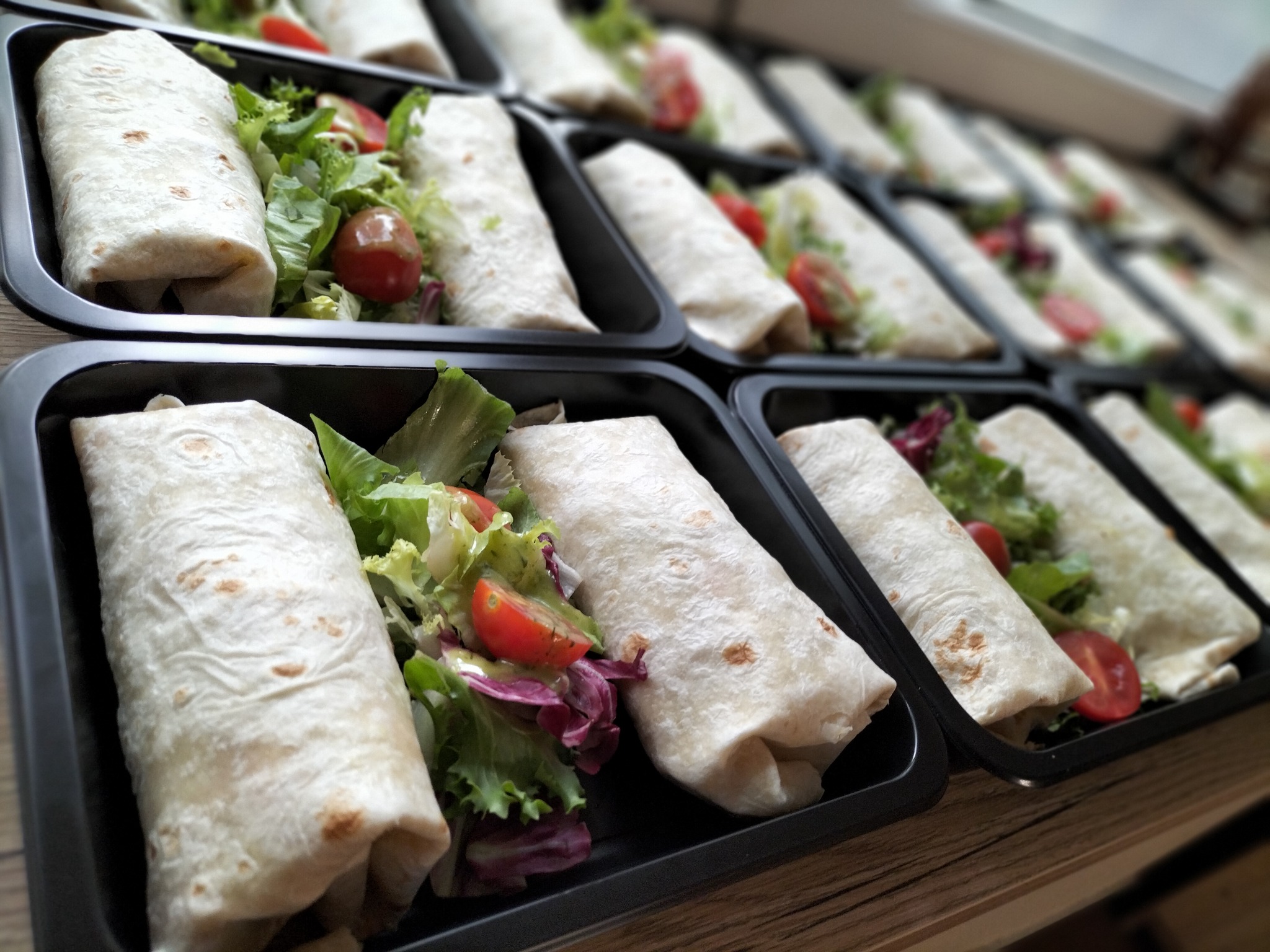 Rząd czarnych pojemników z tortillami nadziewanymi sałatą i pomidorami koktajlowymi, ustawionych ukośnie na drewnianej powierzchni, z rozmytym tłem kolejnych porcji.