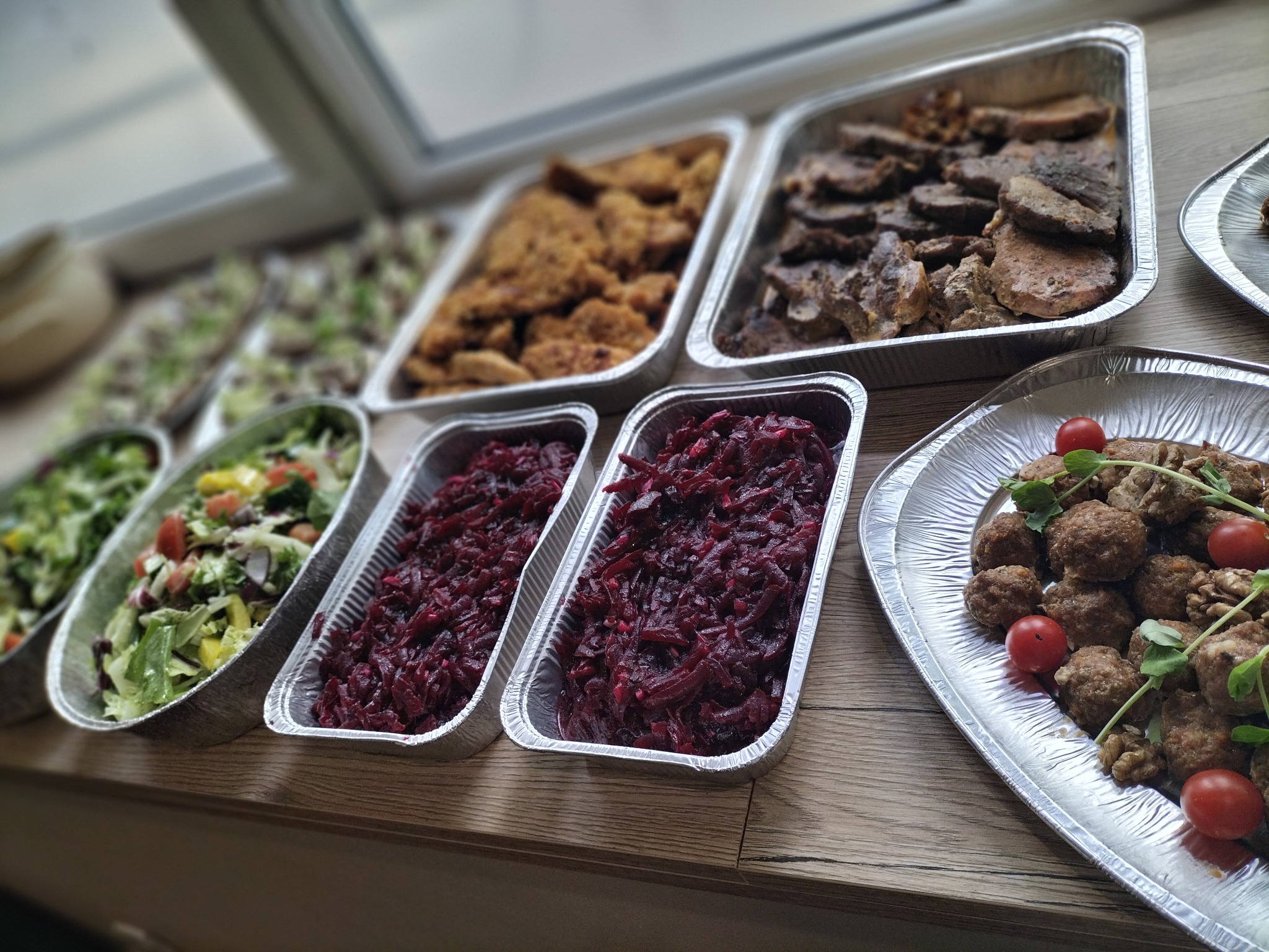 Różnorodne potrawy w aluminiowych tackach ułożone na drewnianym blacie, widoczne sałatki, kotlety schabowe, pieczeń i klopsiki z pomidorkami koktajlowymi.