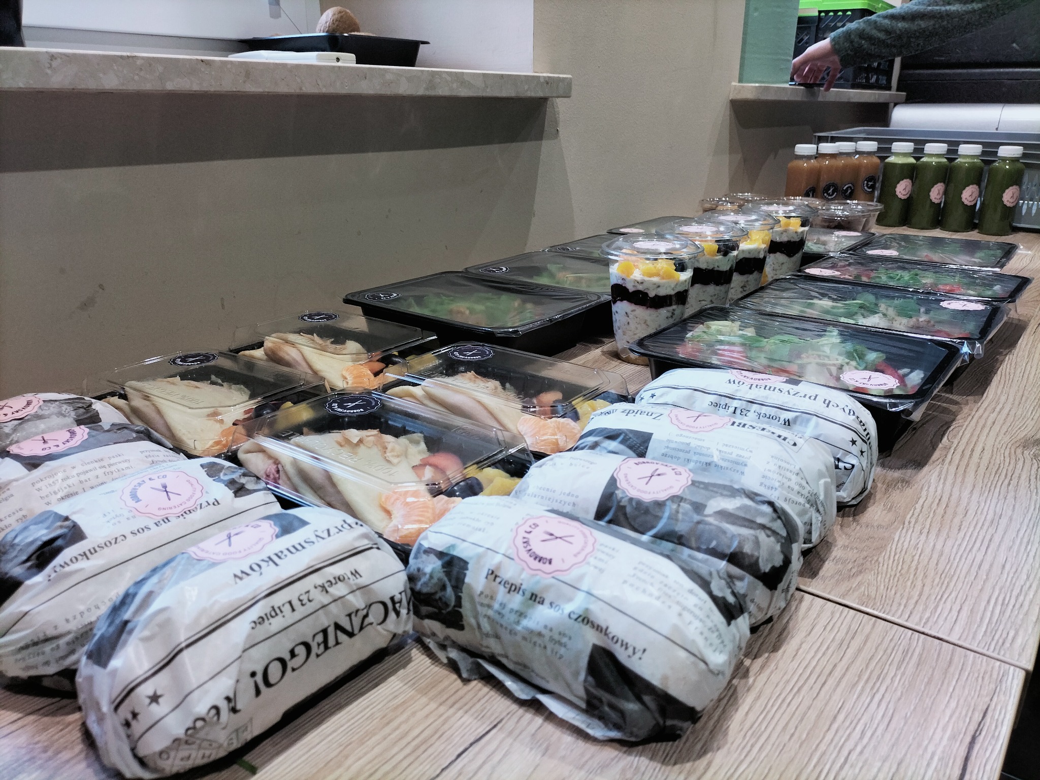 Zestawy cateringowe: sałatki w czarnych pojemnikach, desery z owocami w przezroczystych kubkach i kanapki zawinięte w papier z nadrukowanym tekstem, ułożone rzędami na drewnianym blacie.