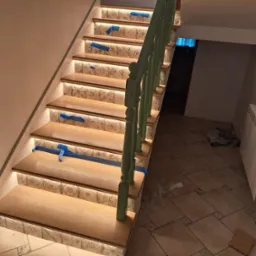 Schody drewniane z podświetleniem LED na stopniach, zabezpieczone niebieską taśmą malarską, z zieloną balustradą i wykończeniem ceramicznym na podstopnicach.