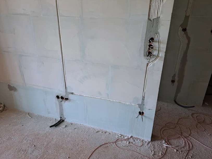 Ściana w trakcie prac wykończeniowych z widocznymi przewodami elektrycznymi, rurkami instalacyjnymi i otworami pod gniazdka, surowy beton na podłodze.