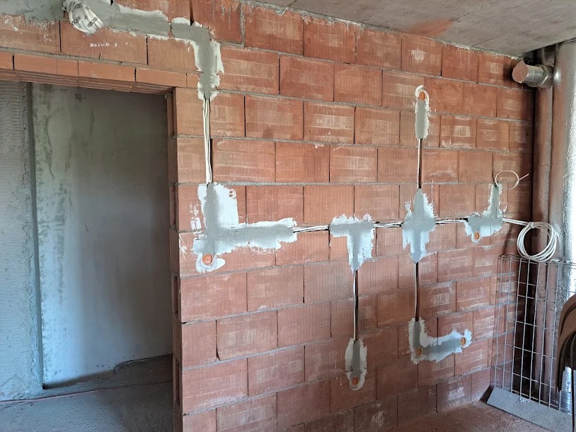 Instalacja przewodów elektrycznych w ścianie z cegły, zabezpieczonych gipsem, widoczne puszki instalacyjne i rury wentylacyjne