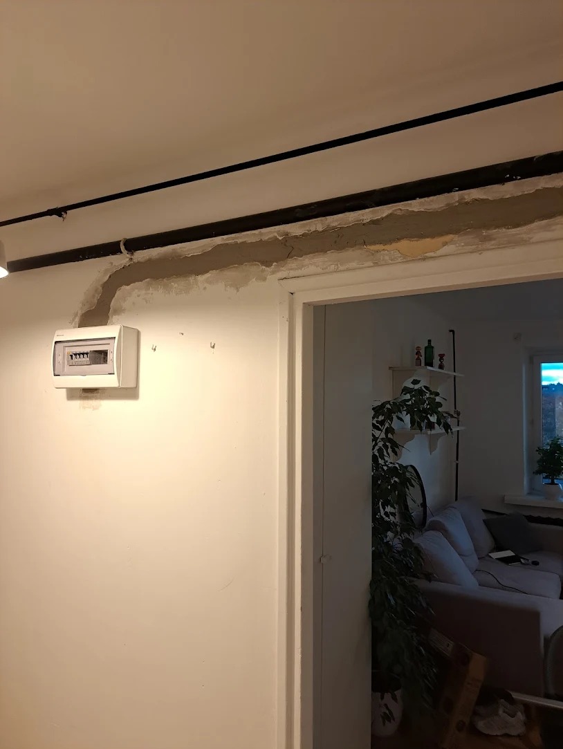 Puszka elektryczna na ścianie, widoczne przewody w listwach na suficie i ścianie, ślady po montażu i szpachlowaniu. W tle widok na salon przez otwarte drzwi.