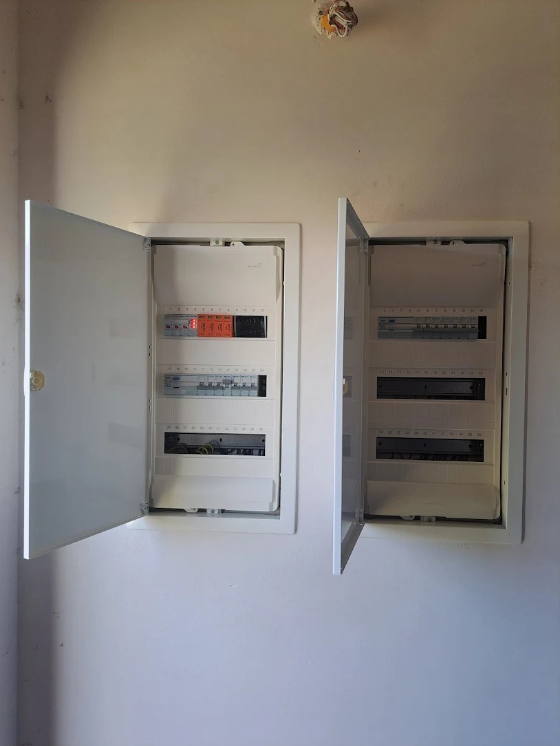 Dwa białe panele elektryczne z otwartymi drzwiczkami na ścianie, widoczne bezpieczniki i okablowanie, surowy stan wykończenia wnętrza.