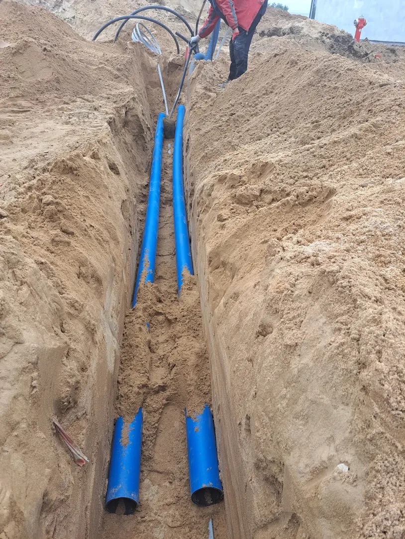 Wykop w piasku z ułożonymi niebieskimi rurami, widoczne kable i fragment osoby w czerwonej kurtce, prace ziemne w toku.