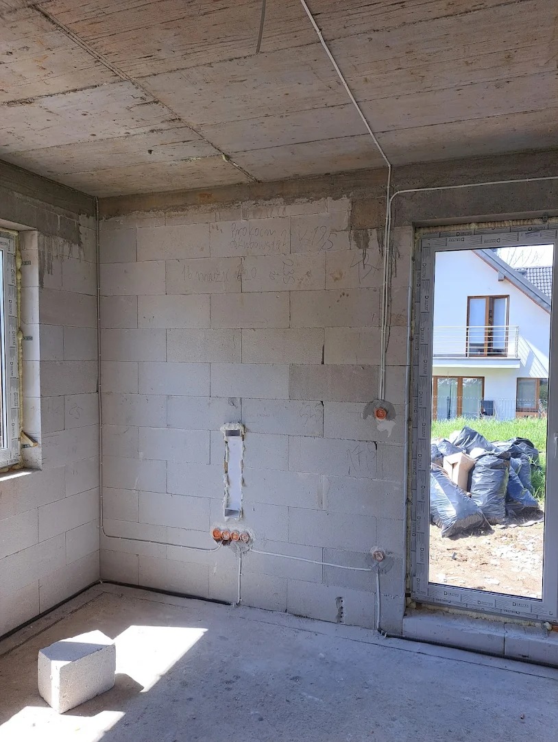 Surowy stan wnętrza z widoczną instalacją elektryczną w ścianach z betonu komórkowego, przygotowaną do dalszych prac wykończeniowych, z oknem na ogród i dom w tle.