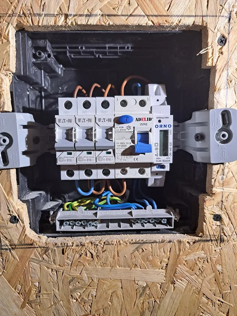 Rozdzielnica elektryczna z zamontowanymi bezpiecznikami EATON i licznikiem ORNO, widoczne przewody w kolorach niebieskim, brązowym i zielono-żółtym, zamocowana w płycie OSB.