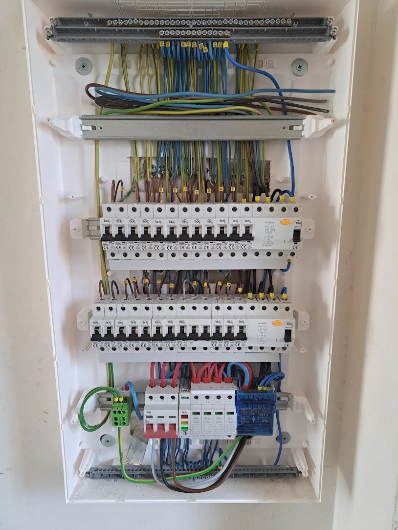 Rozdzielnica elektryczna w białej obudowie z widocznymi bezpiecznikami, przewodami o różnych kolorach oraz listwami zaciskowymi, zamontowana na jasnej ścianie.