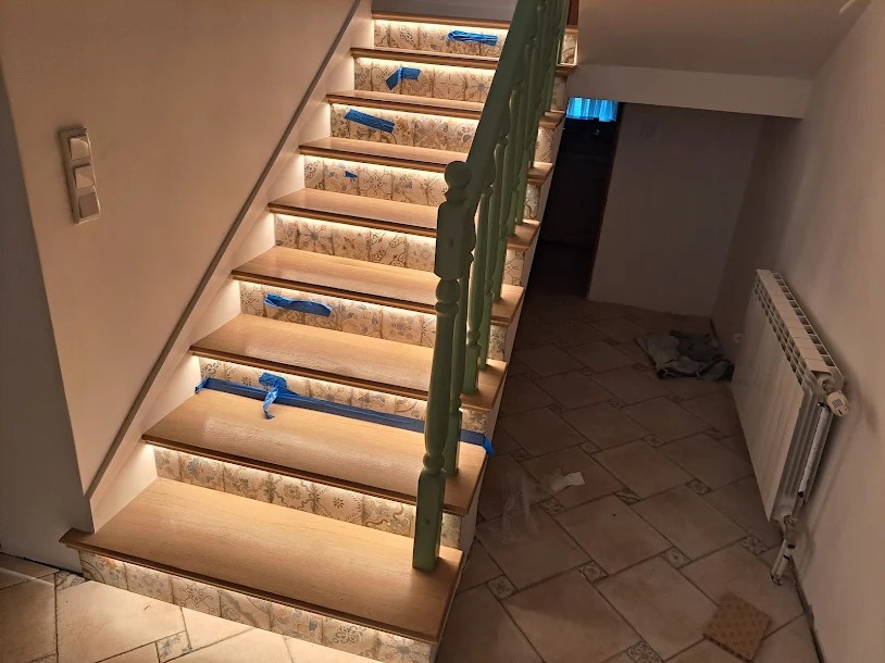 Schody drewniane z podświetleniem LED na stopniach, zabezpieczone niebieską taśmą malarską, z zieloną balustradą i wykończeniem ceramicznym na podstopnicach.