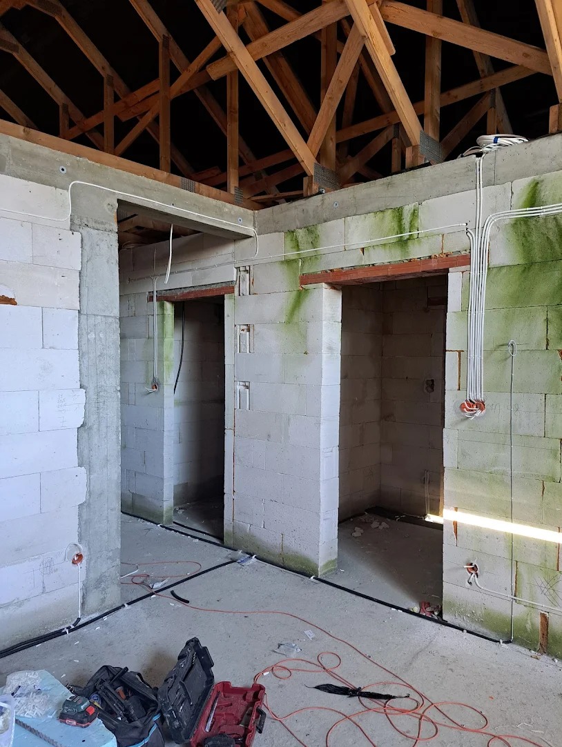 Surowy stan wnętrza z widoczną instalacją elektryczną: kable w peszlach prowadzone po ścianach z betonu komórkowego, widoczne ślady wilgoci, drewniana konstrukcja dachu, narzędzia na podłodze.